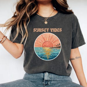 Peut inclure: T-shirt gris foncé avec l'inscription "SUNSET VIBES" au-dessus d'un motif circulaire. Le motif représente un coucher de soleil sur l'océan avec un ciel orange et rose, de l'eau bleue et des silhouettes de palmiers et de personnes.
