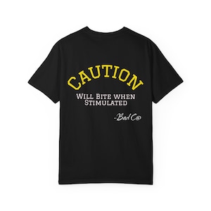 Puede incluir: Camiseta negra con la palabra "CAUTION" en amarillo arqueada sobre "WILL BITE WHEN STIMULATED" y "-Bad Co" en blanco. La camiseta es de cuello redondo.