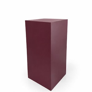 May include: A solid, burgundy-colored rectangular pedestal. The pedestal is a simple, geometric shape with a flat top and a slightly angled base. The surface appears smooth and matte, with no visible texture. The background is plain white.