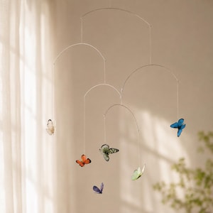 Può includere: Una giostrina sospesa con sottili bracci metallici argentati e arcuati, sostenuti da fili. Sei figure realistiche di farfalle in vari colori, tra cui arancione, blu, verde e bianco, sono fissate alle estremità dei fili a diverse altezze.