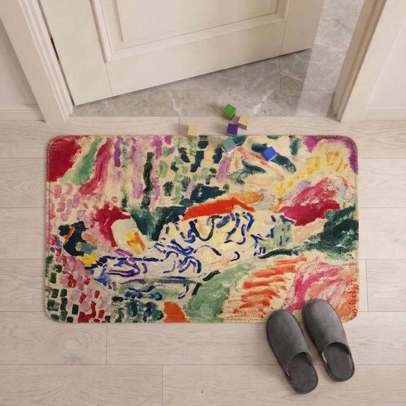 May include: A rectangular floor mat with an abstract design in shades of red, green, yellow, and blue. The mat is placed in front of a doorway, with a pair of gray slippers nearby. Wooden blocks are scattered near the door.