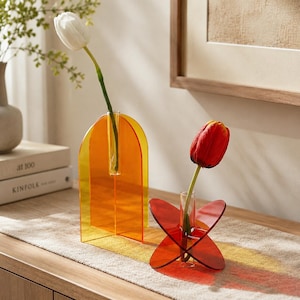 May include: Two acrylic vases with glass inserts. One is a tall, orange, arch-shaped vase holding a white tulip. The other is a smaller, red, cross-shaped vase holding a red tulip. Both vases feature translucent, vibrant colors and geometric designs.