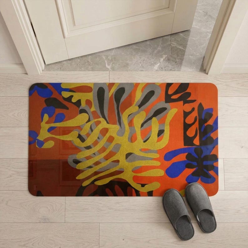 May include: A rectangular doormat with an abstract design in orange, yellow, blue, gray, and black. The mat is placed in front of a white door and next to a pair of gray slippers.