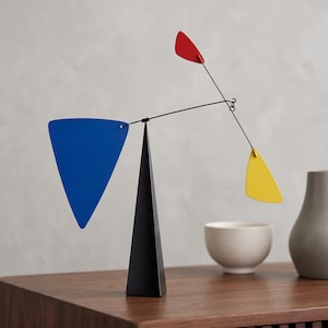 May include: A kinetic sculpture featuring a black triangular base supporting a thin metal rod. Attached to the rod are three flat, geometric shapes in primary colors: a large blue triangle, a small red triangle, and a medium yellow triangle.