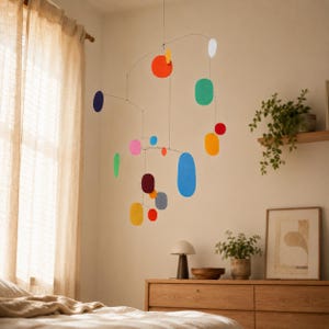 Può includere: Una giostrina sospesa composta da sottili fili metallici e varie forme piatte e colorate, inclusi cerchi e ovali nelle tonalità rosso, arancione, giallo, verde, blu, rosa, viola e grigio, sospesa da un unico punto in equilibrio.