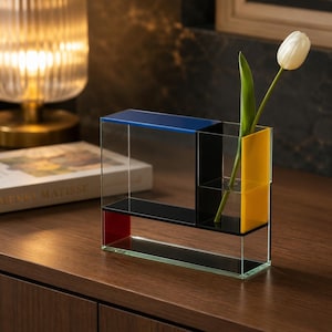 May include: A rectangular clear acrylic vase featuring a geometric design with solid panels in blue, black, yellow, and red. The structure includes a single cylindrical opening for a flower stem, currently holding a white tulip with a green stem.