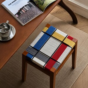 May include: A wooden stool with a rectangular seat featuring a woven pattern of white, blue, yellow, and red threads intersected by thin black lines. The stool has four straight wooden legs and a frame that matches the natural brown wood tone.