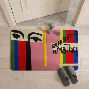 May include: A rectangular doormat with a colorful abstract design featuring a face and the text "EXHIBIT MATISSE". The mat has a pink background with vertical stripes of red, green, blue, and yellow. Gray slippers are placed near the mat.