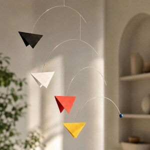 Könnte beinhalten: Ein hängendes Mobile mit vier geometrischen, dreieckigen Elementen in Schwarz, Weiß, Rot und Gelb, die an dünnen, geschwungenen silberfarbenen Metallstäben befestigt sind. Eine kleine blaue Kugel befindet sich am Ende des untersten Metallarms.