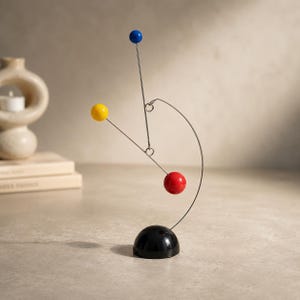 May include: A kinetic sculpture featuring a black semi-spherical base with a thin, curved metal wire extending upwards. Three thin metal rods are suspended from the wire, each tipped with a smooth, solid sphere in primary colours: blue, yellow, and red.