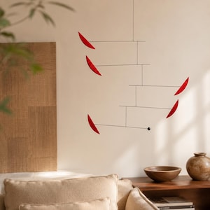 Può includere: Una giostrina sospesa con sottili fili metallici neri collegati in una struttura a livelli. Cinque elementi curvi a forma di foglia rosso brillante sono fissati alle estremità dei fili, bilanciati da una piccola sfera nera sul filo inferiore.