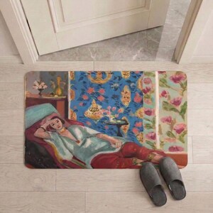 May include: Rectangular doormat featuring a painting of a woman reclining on a chaise lounge. The artwork has a blue, red, and green color palette. Gray slippers are placed near the mat on a light wood floor.