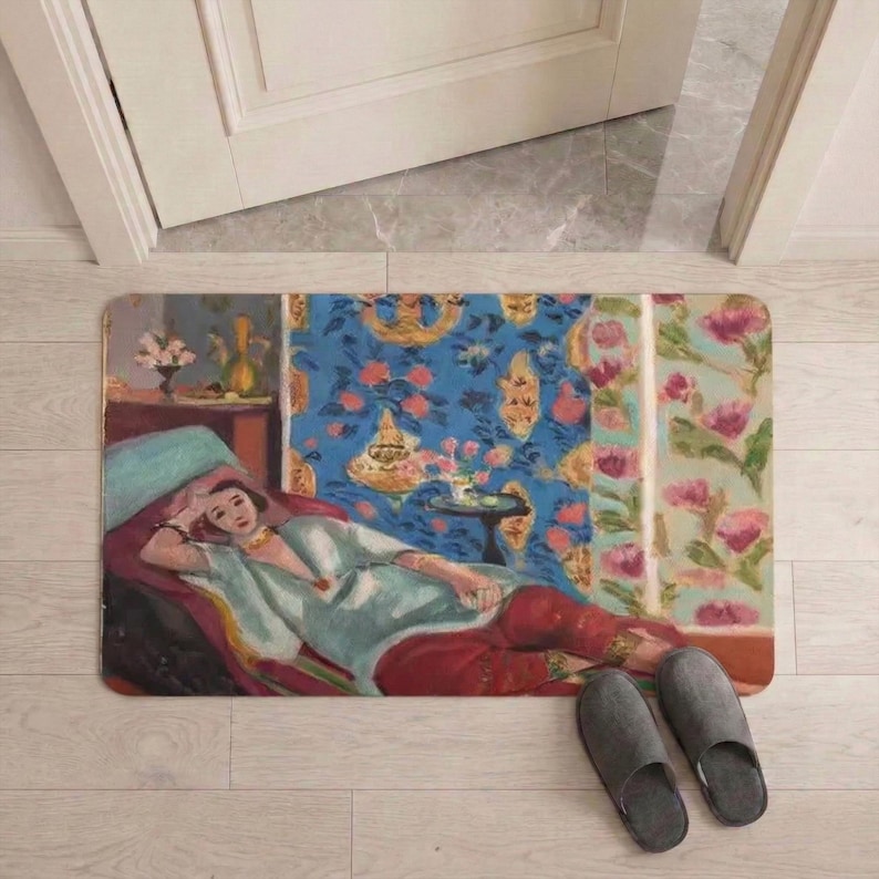 May include: Rectangular doormat featuring a painting of a woman reclining on a chaise lounge. The artwork has a blue, red, and green color palette. Gray slippers are placed near the mat on a light wood floor.