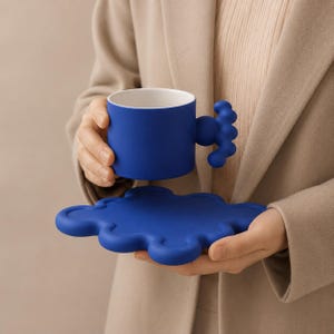 Puede incluir: Un juego de taza y platillo de cerámica en color azul cobalto vibrante. La taza presenta un cuerpo cilíndrico liso con un asa única compuesta por esferas redondeadas apiladas. El platillo tiene un borde ondulado en forma de nube.
