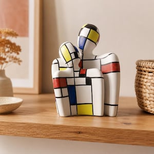 May include: A ceramic sculpture depicting two abstract figures embracing, featuring a white base with a grid pattern of thick black lines and rectangular blocks of primary colors including red, blue, and yellow across the entire surface.