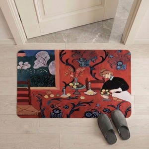 May include: Rectangular doormat featuring a vibrant painting of a woman at a table with fruit and flowers. The mat has a red background with blue floral patterns and a scene of a landscape. Gray slippers are in the foreground.