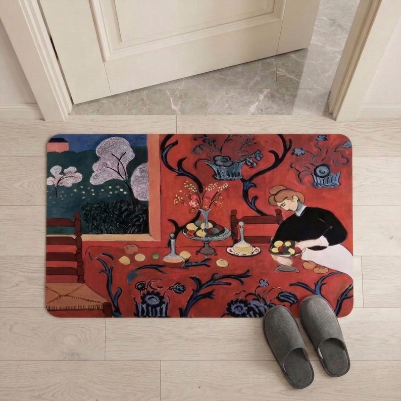 May include: Rectangular doormat featuring a vibrant painting of a woman at a table with fruit and flowers. The mat has a red background with blue floral patterns and a scene of a landscape. Gray slippers are in the foreground.