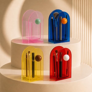 Op de afbeelding: Vier doorschijnende acryl organizers met gebogen bovenkanten, elk voorzien van een klein bolvormig accent. De organizers zijn roze, blauw, geel en rood en bevatten diverse make-up kwasten, potloden en mascara voor opslag.