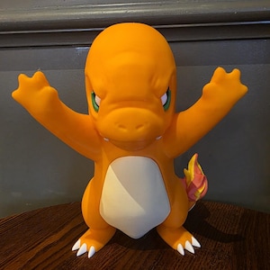 May include: A bright orange Charmander figurine with a cream-colored belly, green eyes, and white claws. The character is depicted in a standing pose with both arms raised upward, and a small flame with yellow and pink accents on its tail.