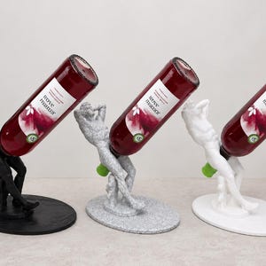 May include: Three wine bottle holders shaped like human figures in black, speckled grey, and white. Each figure is seated on an oval base, holding a bottle of Wove Manor red wine at an angle. The figures have a muscular build and are posed with arms behind their heads, supporting the bottle neck with their legs.