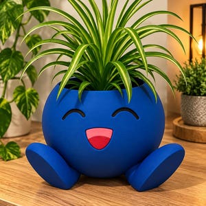 May include: A round, bright blue planter shaped like a character with two feet, a wide smiling mouth with a pink tongue, and two curved black eyes. A green and white spider plant grows out of the top of the blue, smooth-textured container.
