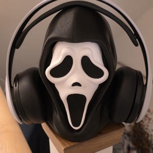 Può includere: Una maschera di Ghostface in bianco e nero con un cappuccio nero, che indossa cuffie nere e bianche. La maschera, con il suo iconico volto bianco e le orbite e la bocca nere, è al centro. Le cuffie sono posizionate attorno alla maschera.