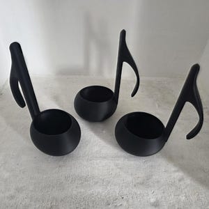 May include: Three black musical note-shaped planters. Each planter has a round bowl and a curved stem, resembling a musical note. The planters are made of a matte black material and are arranged on a light grey surface.