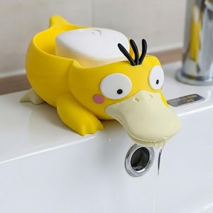 Peut inclure: Un porte-savon jaune vif en forme de canard de dessin animé avec un large bec crème, de grands yeux blancs, des touffes de cheveux noirs et des taches roses sur les joues. Le canard porte un pain de savon blanc rectangulaire sur son dos.