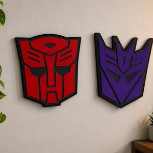 May include: Two wall-mounted decorative plaques featuring the Autobot and Decepticon logos. The Autobot emblem is red with black outlines, while the Decepticon emblem is purple with black outlines. Both symbols have a layered, three-dimensional design.