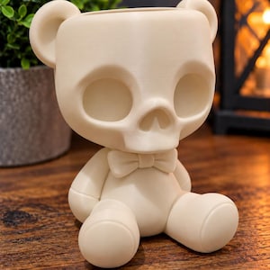 May include: A cream-coloured planter shaped like a seated teddy bear with a skull-like face. The figure features large round ears, hollow eye sockets, a small nose cavity, and a bow tie at the neck. The smooth, matte-finished object is hollow at the top.