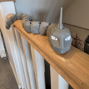 May include: A grey, segmented sculpture of an Onix, a Pokémon character, on a wooden handrail. The sculpture has a detailed head with white eyes and a horn, and a body made of connected rock-like segments.