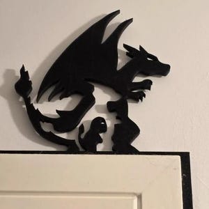 May include: A black dragon-themed decorative piece, possibly a door or window frame topper. The silhouette features a dragon with large wings, a flame, and two smaller figures. The piece is mounted on a white door frame.