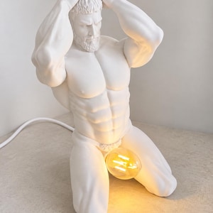 May include: A white matte sculpture lamp depicting a muscular figure kneeling with hands behind the head. The figure features a detailed beard and torso, with a glowing amber-toned Edison-style light bulb positioned at the pelvic area.