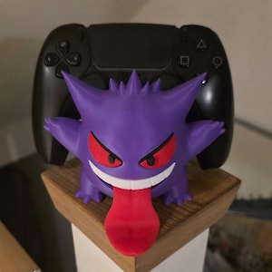 Puede incluir: Una figura del personaje Gengar de color morado con ojos rojos, boca blanca y una larga lengua roja que cuelga. La figura está colocada frente a un mando de videojuegos negro, sirviendo como soporte para el dispositivo sobre una superficie de madera.