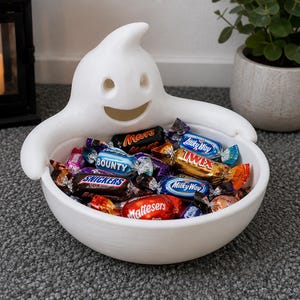 Könnte beinhalten: Eine weiße Keramikschale in Form eines Geistes mit lächelndem Gesicht und ausgestreckten Armen, gefüllt mit einer Auswahl an einzeln verpackten Schokoriegeln, darunter Snickers, Mars, Bounty, Twix, Milky Way und Maltesers.