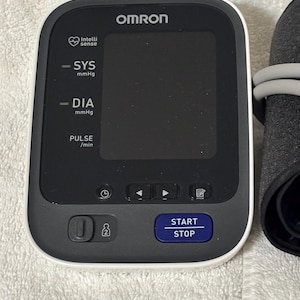 May include: An Omron blood pressure monitor with a white frame and a black digital display screen. The device features labels for SYS mmHg, DIA mmHg, and PULSE /min, along with a blue START STOP button and icons for settings and user selection.