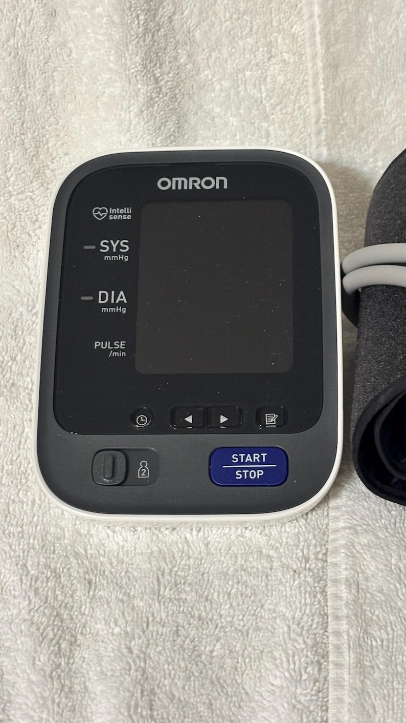 May include: An Omron blood pressure monitor with a white frame and a black digital display screen. The device features labels for SYS mmHg, DIA mmHg, and PULSE /min, along with a blue START STOP button and icons for settings and user selection.