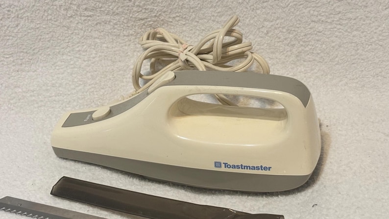Puede incluir: Cuchillo el&eacute;ctrico para trinchar crema y gris con mango y cable. El cuchillo tiene una base y un mango grises, con la marca "Toastmaster" impresa en el lateral. Se ven dos cuchillas.