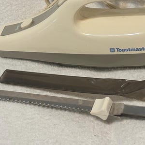Puede incluir: Un cuchillo el&eacute;ctrico para trinchar Toastmaster de color crema con una hoja dentada y una funda protectora. El cuchillo tiene un mango blanco y una base gris. El nombre de la marca est&aacute; impreso en el lateral.