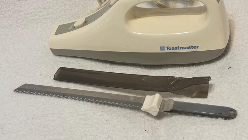 Puede incluir: Un cuchillo el&eacute;ctrico para trinchar Toastmaster de color crema con una hoja dentada y una funda protectora. El cuchillo tiene un mango blanco y una base gris. El nombre de la marca est&aacute; impreso en el lateral.