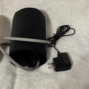 May include: A black blood pressure monitor cuff with a gray rubber tube wrapped around it. A black power adapter with a two-prong plug and a thin black cord with a small circular connector is positioned next to the cuff on a textured surface.