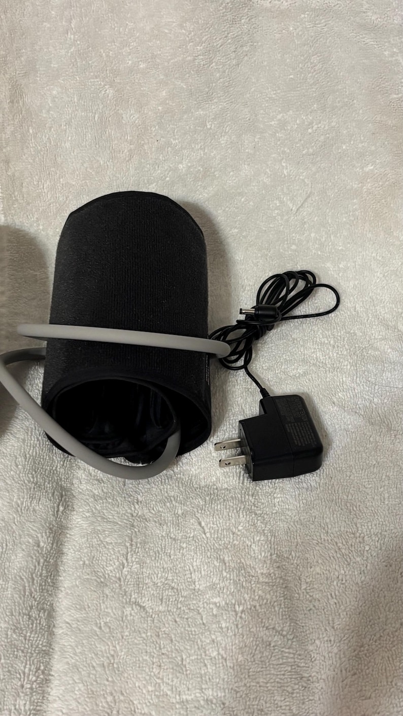 May include: A black blood pressure monitor cuff with a gray rubber tube wrapped around it. A black power adapter with a two-prong plug and a thin black cord with a small circular connector is positioned next to the cuff on a textured surface.