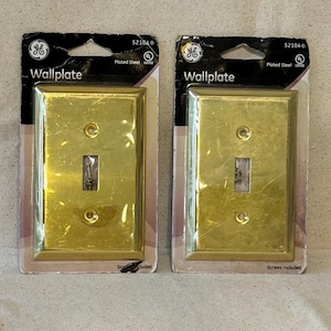 Puede incluir: Dos placas de pared doradas en su embalaje original. Cada placa de pared tiene forma rectangular con una abertura para un solo interruptor y dos orificios para tornillos. El embalaje incluye el texto "Wallplate" y el logotipo de GE.
