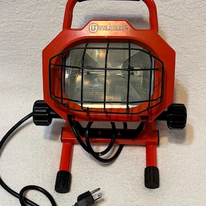 May include: Red UTILITECH work light with a black handle, protective cage, and adjustable knobs. The light has a black power cord with a standard US plug. The light is designed for work or utility purposes.