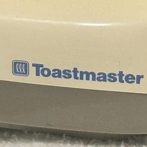 Puede incluir: Primer plano de un electrodom&eacute;stico color crema con el texto azul "Toastmaster". El nombre de la marca se muestra de forma destacada en la parte frontal del electrodom&eacute;stico. El electrodom&eacute;stico tiene una forma redondeada.