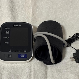 May include: An Omron blood pressure monitor with a white and black digital display unit, a black arm cuff with a gray air tube, and a black power adapter. The display features the text OMRON, SYS, DIA, PULSE, and a blue START STOP button.