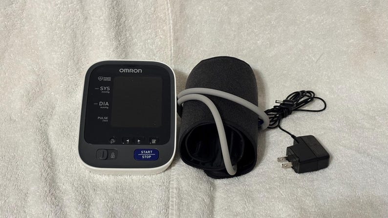 May include: An Omron blood pressure monitor with a white and black digital display unit, a black arm cuff with a gray air tube, and a black power adapter. The display features the text OMRON, SYS, DIA, PULSE, and a blue START STOP button.