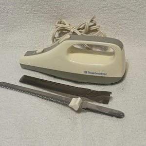 Puede incluir: Un cuchillo el&eacute;ctrico para trinchar de color blanquecino y gris con una hoja dentada y una funda protectora. El cuchillo tiene un mango con un lazo y la marca "Toastmaster" est&aacute; impresa en el lateral.