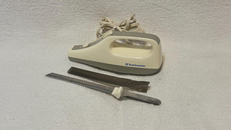 Puede incluir: Un cuchillo el&eacute;ctrico para trinchar de color blanquecino y gris con una hoja dentada y una funda protectora. El cuchillo tiene un mango con un lazo y la marca "Toastmaster" est&aacute; impresa en el lateral.