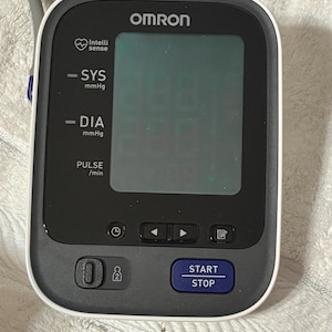 May include: An Omron blood pressure monitor with a black and white casing, a large digital display screen, and a blue Start/Stop button. The device features labels for SYS mmHg, DIA mmHg, and PULSE/min, along with navigation and memory buttons.